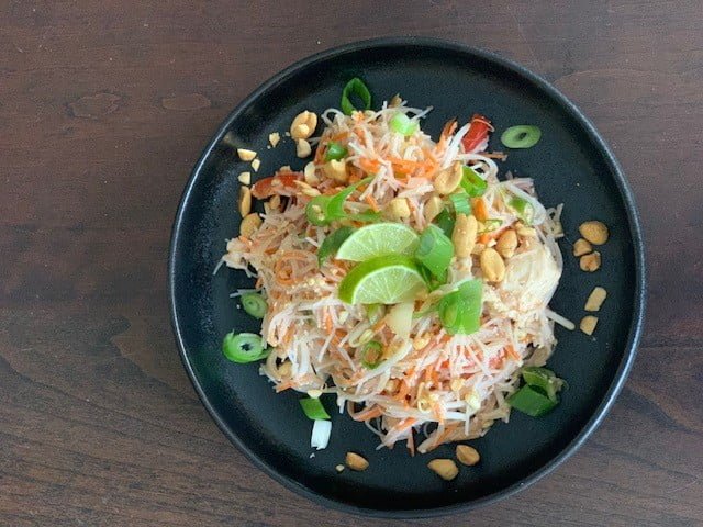 Een zwart bord met opstaande rand met daarop de Pad Thai met Dzjing> Er liggen een paar partjes limoen op ter garnering