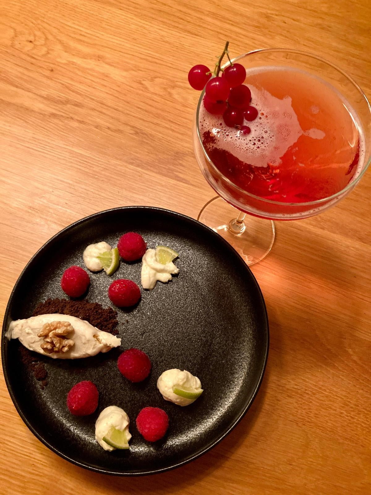 Een zwart bord met daarop een walnotenparfait, crumble, limoenmousse met frambozen en een champagnecoupe met VeldFlora mousserende thee cuvée met een trosje rode bessen over de rand.