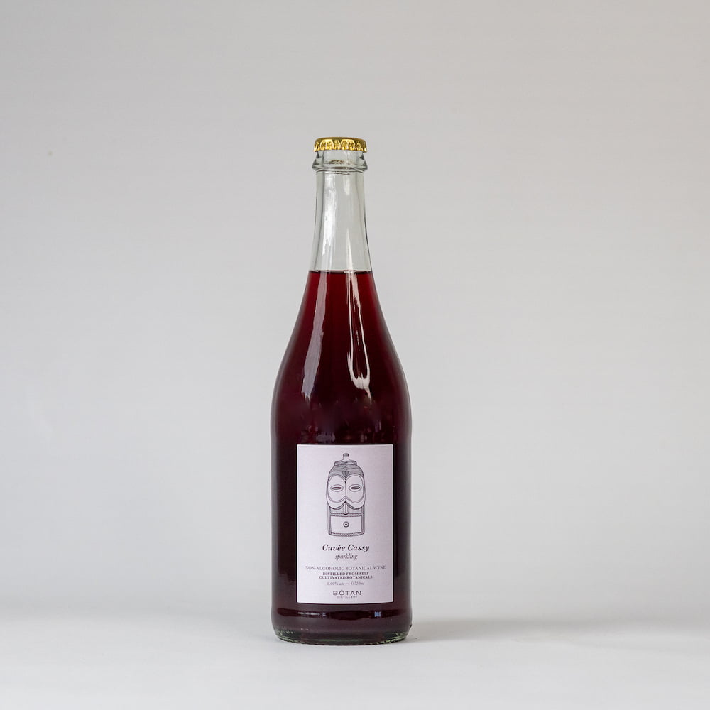 Een fles Cuvée Cassy Botanical Wyne. Een lichtroze etiket met een foto van een kleine totempaal, Cuvée Cassy is rood van kleur en de fles wordt afgesloten met een kroonkurk