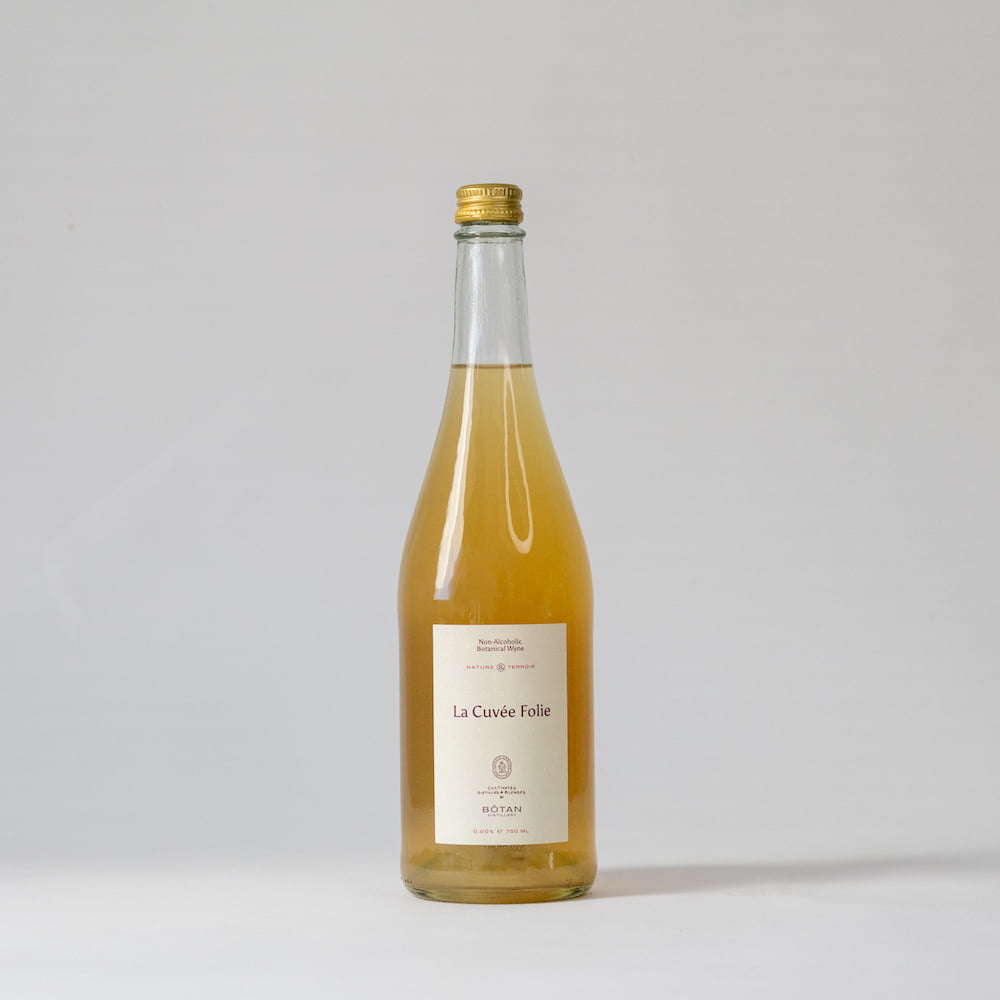 Een fles La Cuvée Folie met een licht etiket. De drank zelf is goudgeel van kleur en de fles is afgesloten emt een kroonkurk