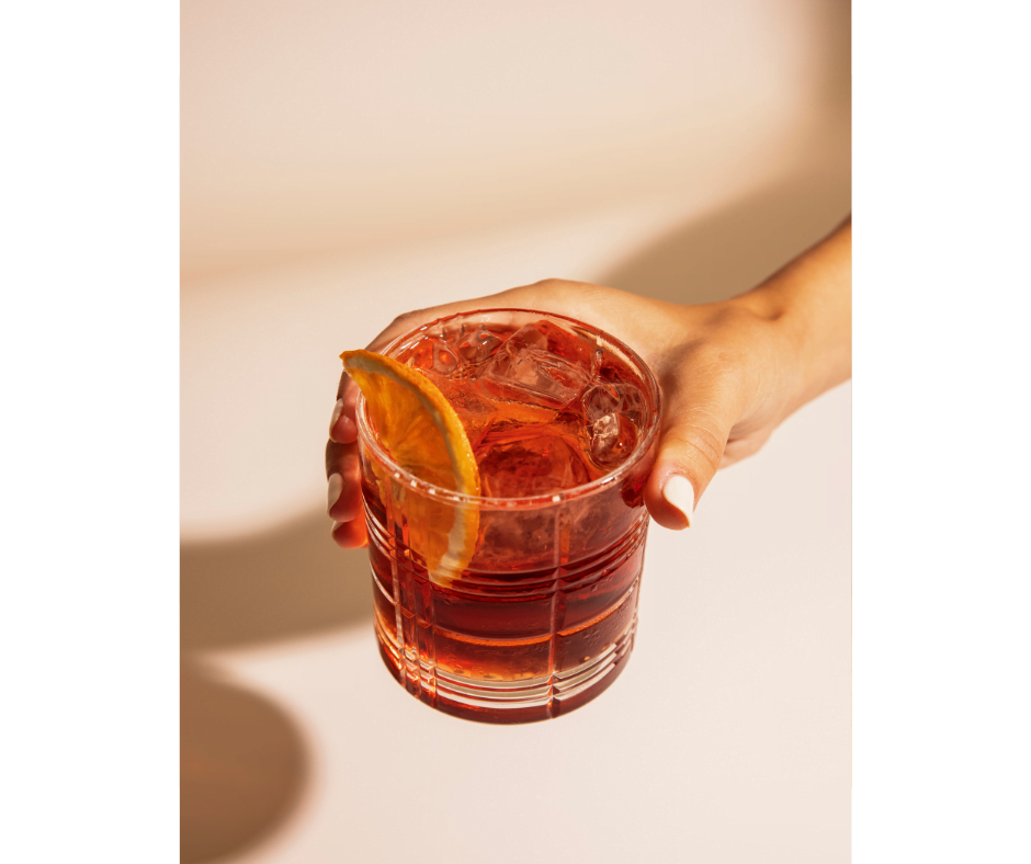 Sunshine Reggae alcoholvrije cocktail. Een hand heeft een tumblerglas vast, gevuld met de rood gekleurde sunshine reggae cocktail, ijsklontjes en afgewerkt met een schijfje sinaasappel