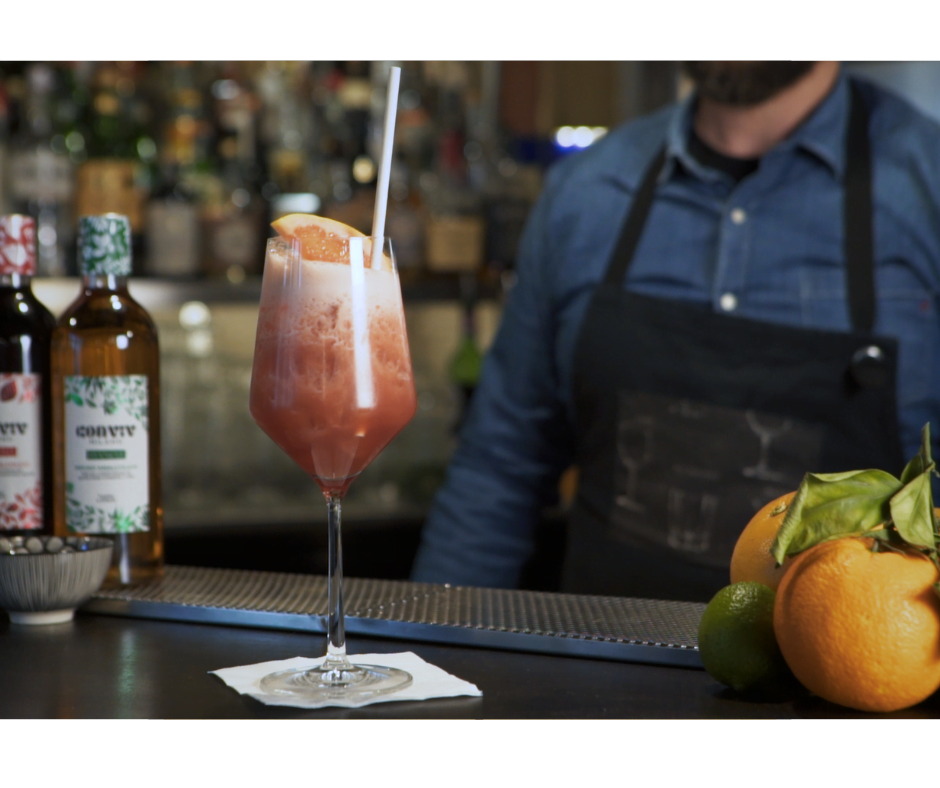 Op de bar staat een schuimige cocktail met een mooie roze kleur, gegarneerd met een schijfje sinaasappel en een rietje. Achter de bar staat barman Pier